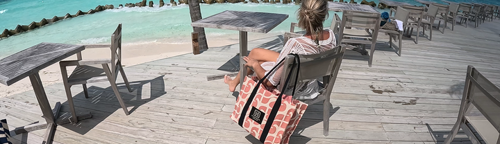 Roze Amora Tote Bag hangt aan een houten stoel terwijl een model zit op een boulevard met uitzicht op tropisch water