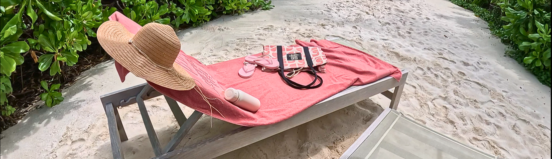 Strandset op een strandbedje in het zand met een roze strandlaken een roze strandtas, roze slippers een drinkbeker om je drankje koud te houden