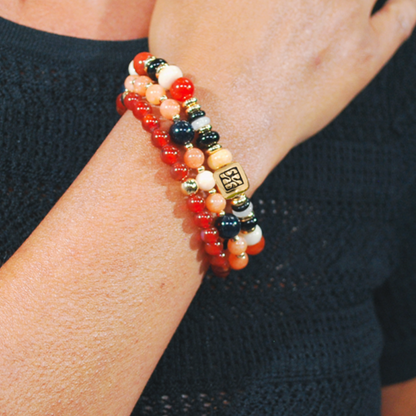 Model draagt de Sunset Mix armbandenset van SummerGear met rode, oranje en zwarte kralen en goudkleurige details bij een zwarte top.